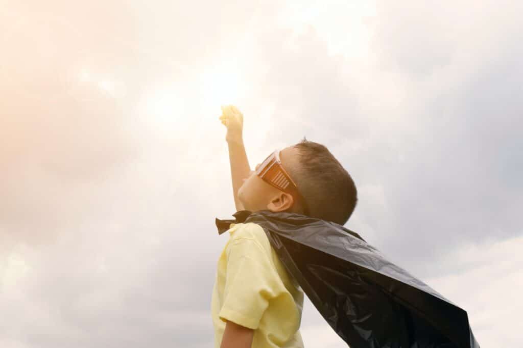Criança com capa de super-herói com o braço para cima fingindo que está voando com seus super poderes.